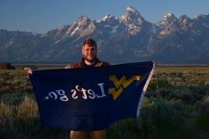 Joshua Carder is holding a banner with the WV logo and the words "Let's go." while standing in a large field with rocky mountains far behind him